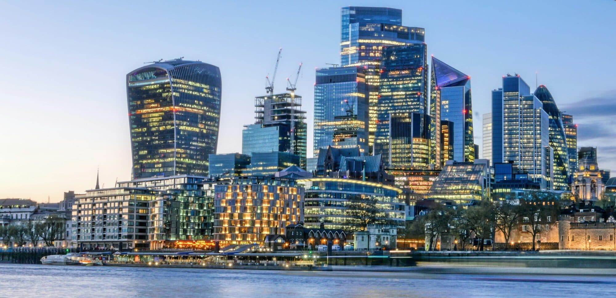 London financial district skyline at dusk with illuminated skyscrapers and River Thames in the foreground London financial district skyline at dusk with illuminated skyscrapers and River Thames in the foreground